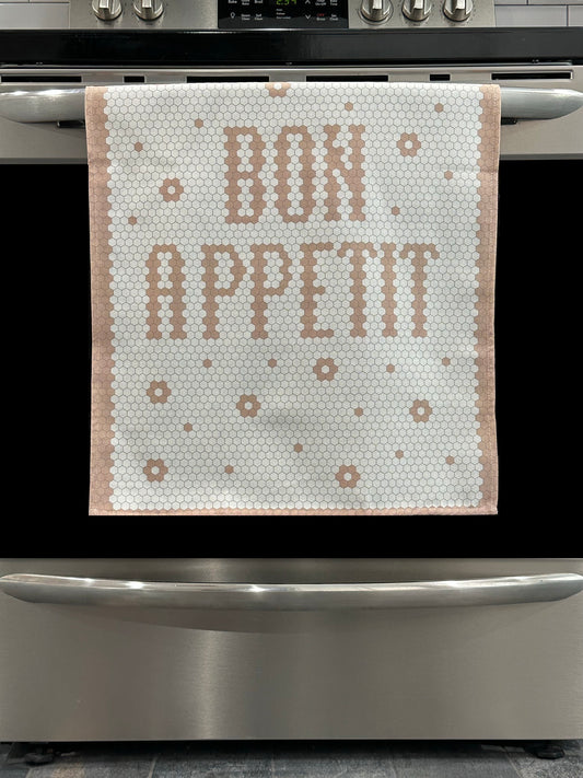 Kitchen Towel in white and blush featuring a "Bon Appetit" design. Pictured here hanging on a stove.