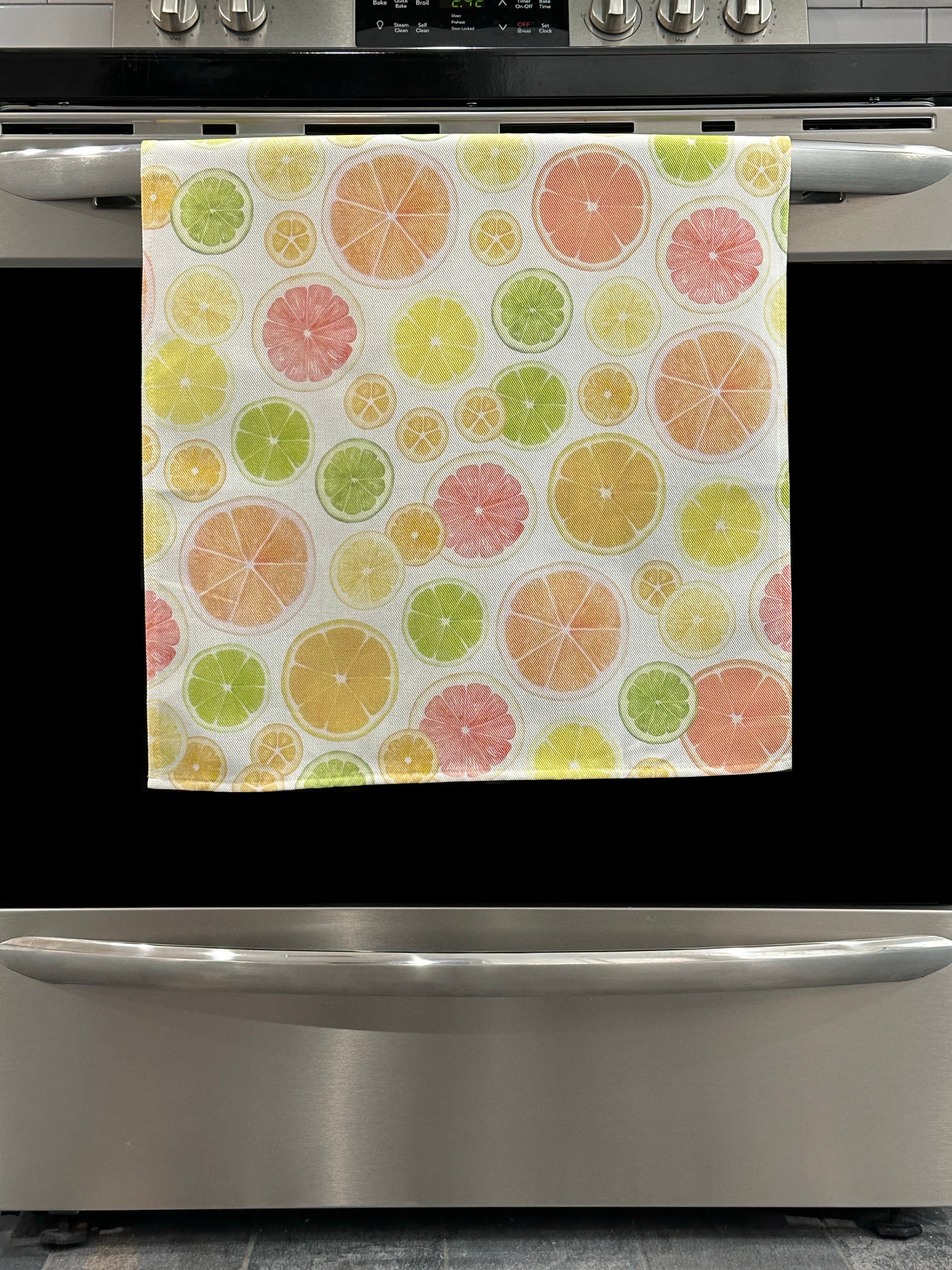 Kitchen Towel featuring a multicolor citrus fruit design. Pictured here hanging on a stove.