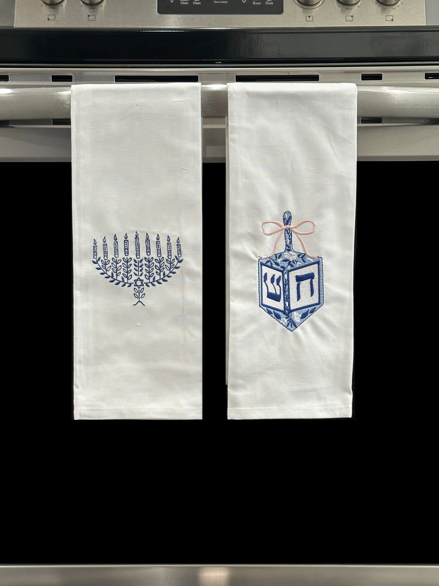 Two Hanukkah Kitchen Towels. On the left, a kitchen towel featuring a blue embroidered menorah. On the right, a blue and pink embroidered dreidel. 